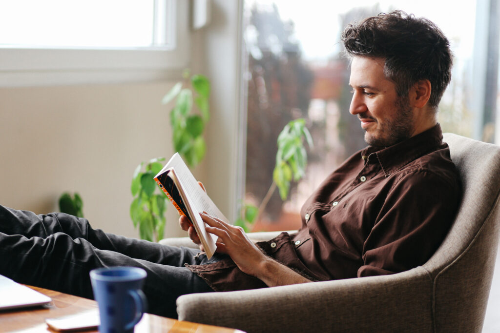 Vintage toned portrait of a young man sitting at the table in the bright and beautiful apartment on the high floor in Belgrade, Serbia. He is wearing casual, everyday clothes, a brown shirt, reading a book and having a cup of coffee as well.